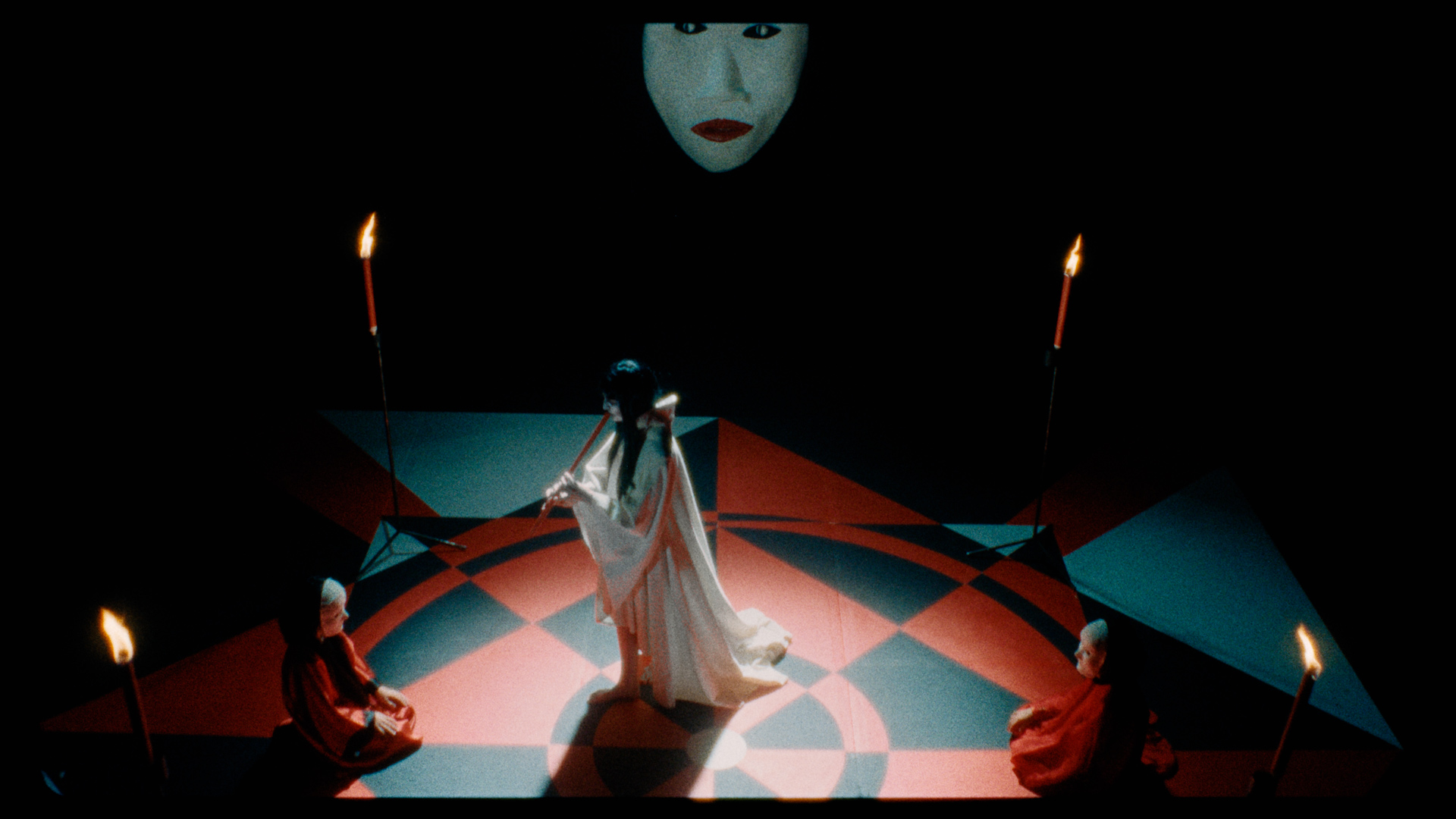 A mask-wearing woman playing the flute, surrounded by kneeling figures in white masks.