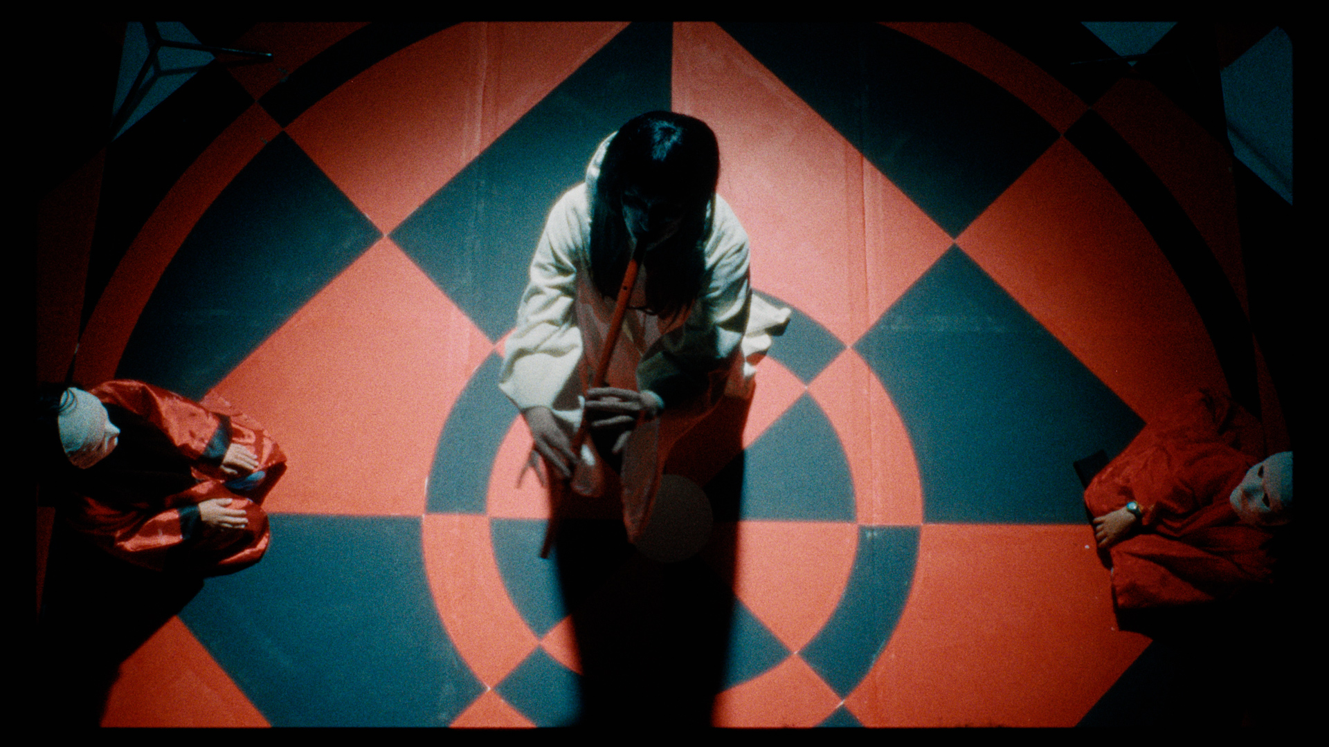 A mask-wearing woman playing the flute, surrounded by kneeling figures in white masks.